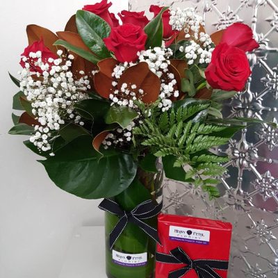 A tall vase of blooming red roses, baby's breath and green foliage with a wrapped box next to it