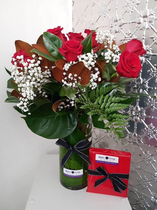 A tall vase of blooming red roses, baby's breath and green foliage with a wrapped box next to it