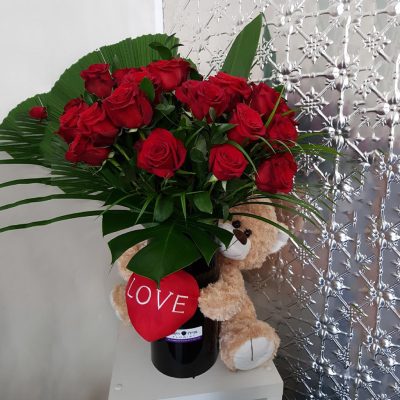 3 dozen red roses with green foliage in a large black vase with a teddy holding a heart wrapped around it.
