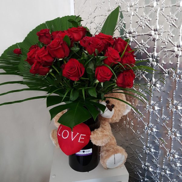 3 dozen red roses with green foliage in a large black vase with a teddy holding a heart wrapped around it.